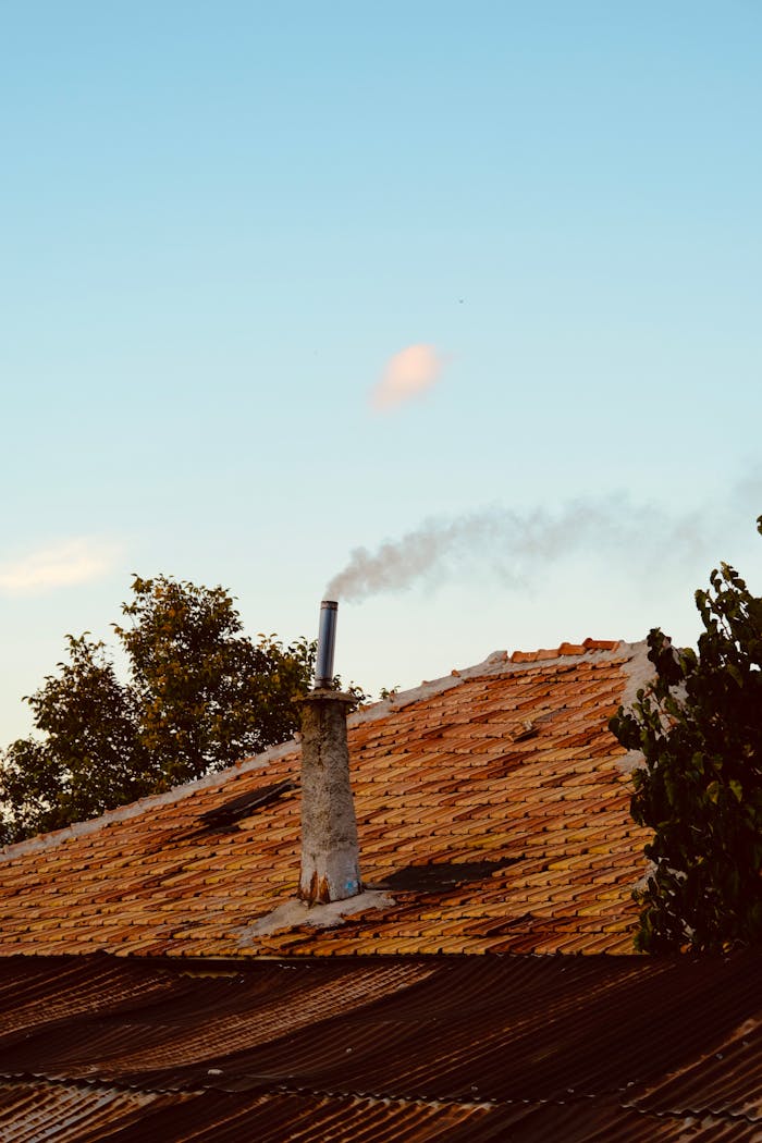A rustic tiled roof with a smoking chimney set against a clear blue sky.